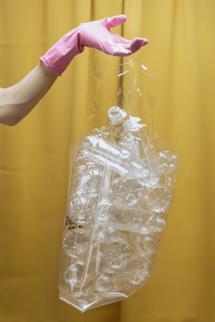A person wearing a light pink rubber glove is holding a large, transparent plastic bag filled with crumpled clear plastic bottles and containers. The bag is suspended in mid-air against a yellow curta