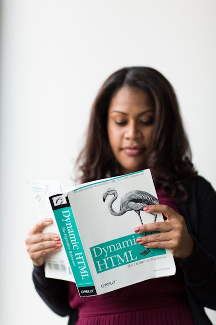 A woman with dark, shoulder-length hair, dressed in a black blazer and burgundy top, is holding a book titled 'Dynamic HTML' with a cover featuring an illustration of a flamingo. She is positioned aga