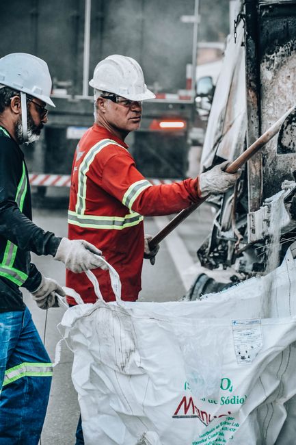 Two construction workers, both wearing protective white helmets and safety gloves, are in the process of removing debris from the burnt-out front section of a damaged vehicle on a city street. The wor